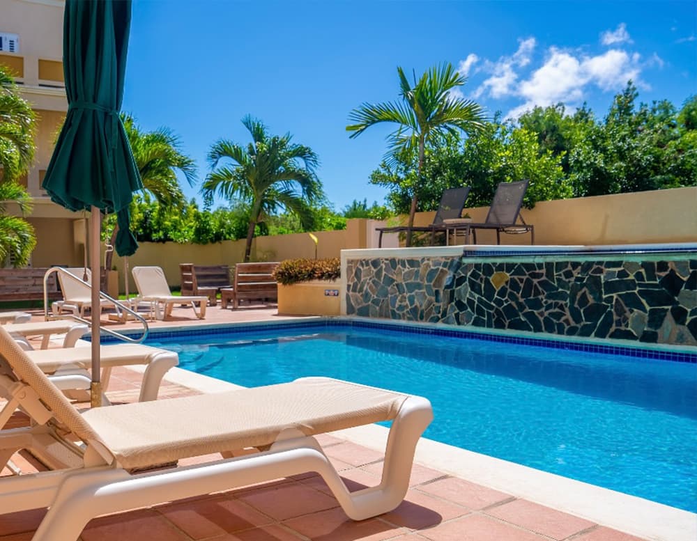 A sunny pool area with lounge chairs, surrounded by tropical plants and a clear blue sky.